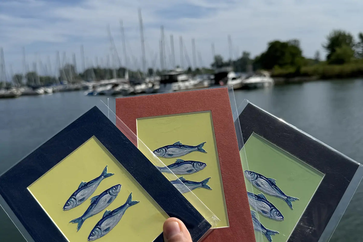 A hand holding three sardine mini art frames in front of a Lake Ontario marina